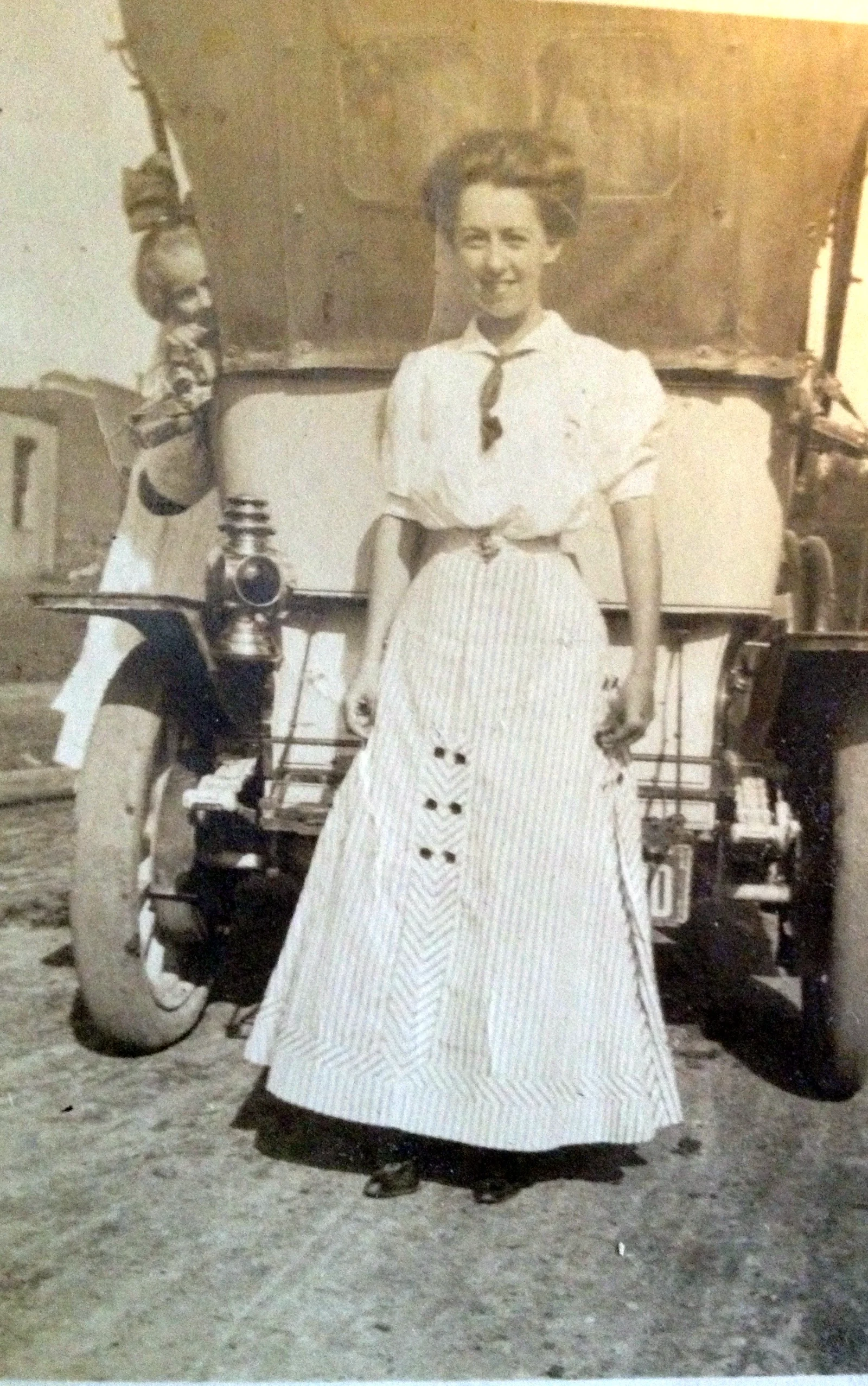 Vintage photograph of Luta Lee Helton Shrum standing in front of an early automobile, wearing a long striped dress, early 1900s.