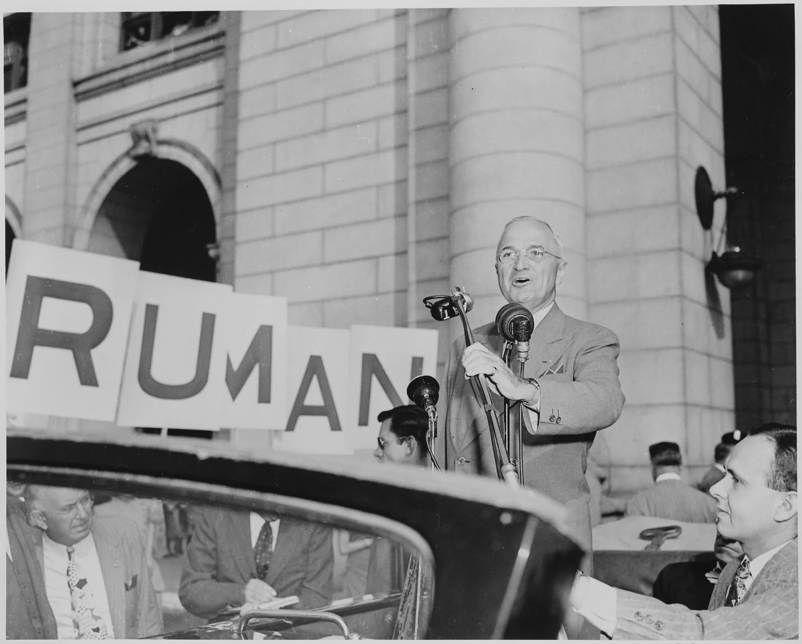 President Harry S. Truman speaks into microphones from an open car during the 1948 presidential campaign, with a large Truman sign behind him.