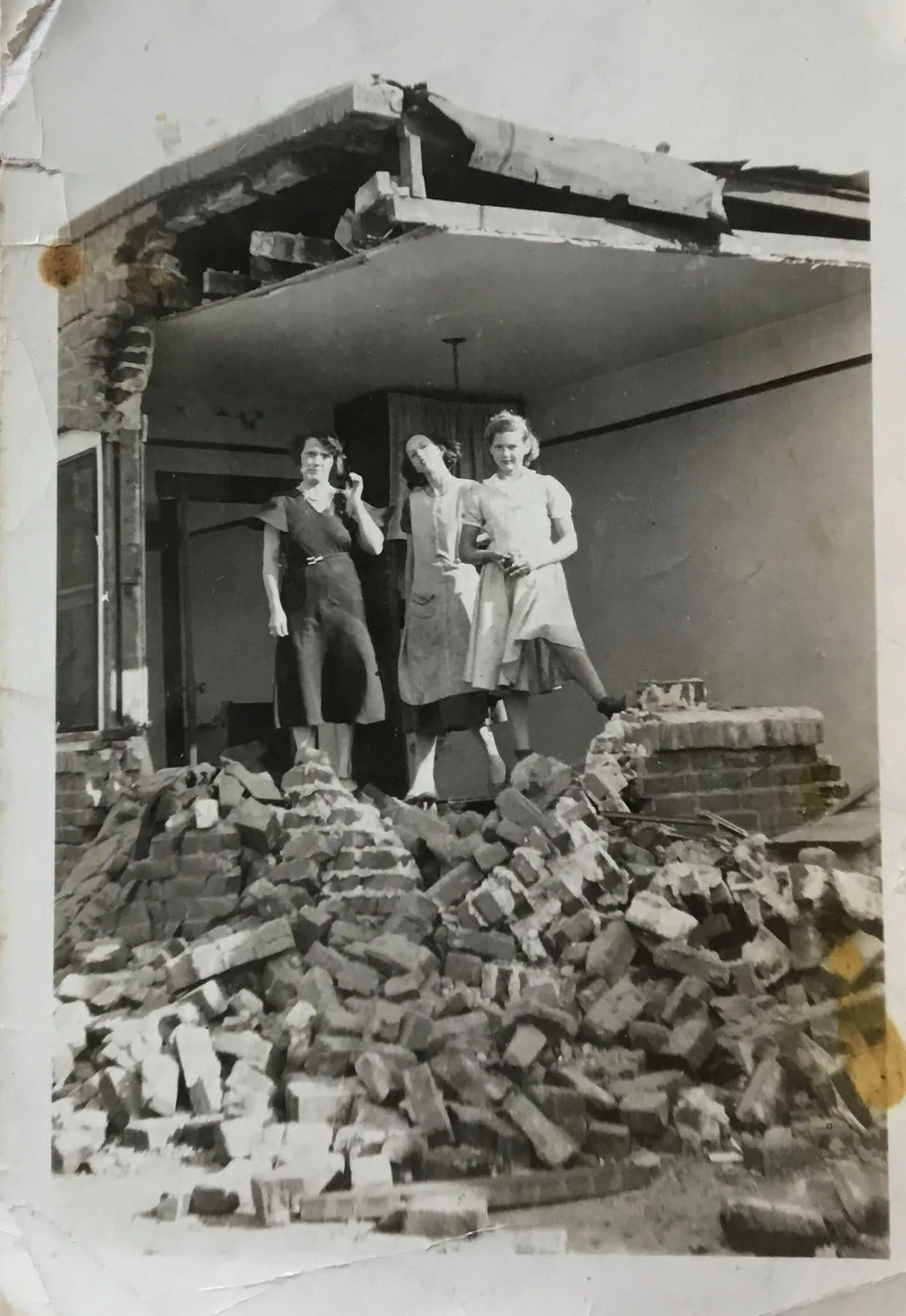 Three women identified as Mamie, Evelyn, and Dorothy Braden stand atop a large pile of rubble inside their collapsed Long Beach home following the March 10, 1933 earthquake. A damaged ceiling and broken brick walls are visible behind them.