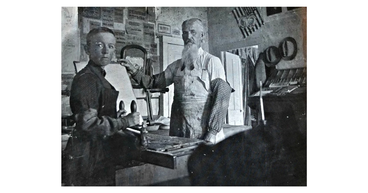 Black and white photograph of two men working in an early newspaper print shop, with a typesetting tray on the workbench in front of them, newspaper clippings and an American flag visible on the walls in the background, late 1800s.