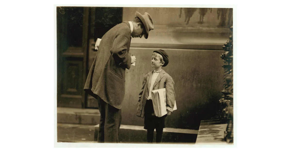 Sepia-toned photograph of a man leaning down to speak with a young newsboy holding a stack of newspapers on a city street, early 1900s.